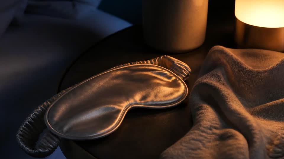 A dim bedside scene with a silk sleep mask, folded throw and a low glowing lamp